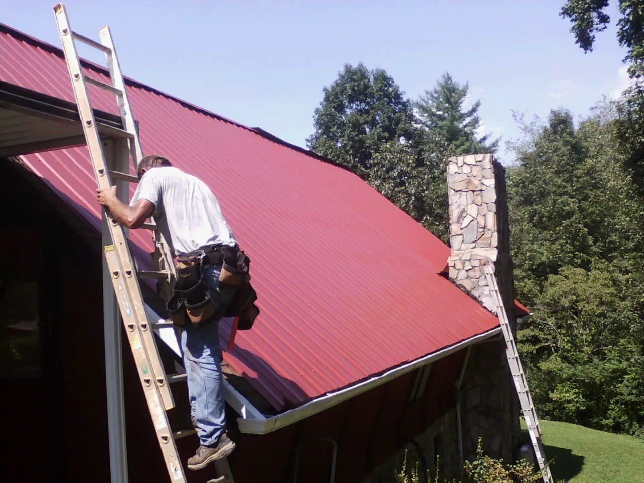 Roofer on a ladder beside a red metal roof and stone chimney — Soffit & Fascia Repair by Roofing Brothers in Wausaukee